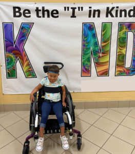 Dianne's daughter in wheelchair sitting in front of a decorative banner that reads "Be the I in Kind"